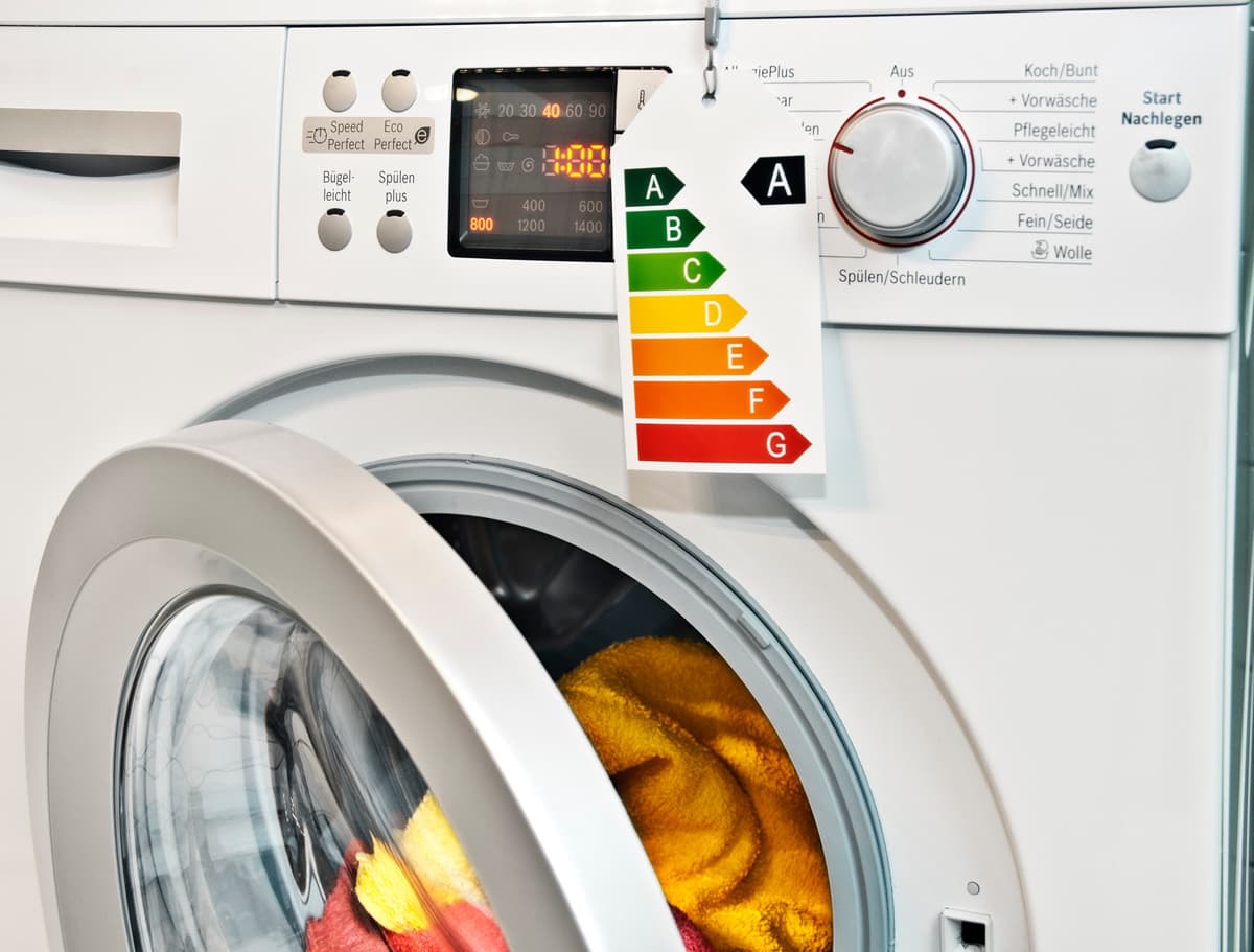 Eine moderne Frontlader-Waschmaschine mit transparenter Tür, durch die bunte Kleidungsstücke sichtbar sind. An der Maschine hängt ein Energielabel mit einer Skala von A bis G, wobei die höchste Effizienzklasse A hervorgehoben ist. Das Bedienfeld zeigt ver