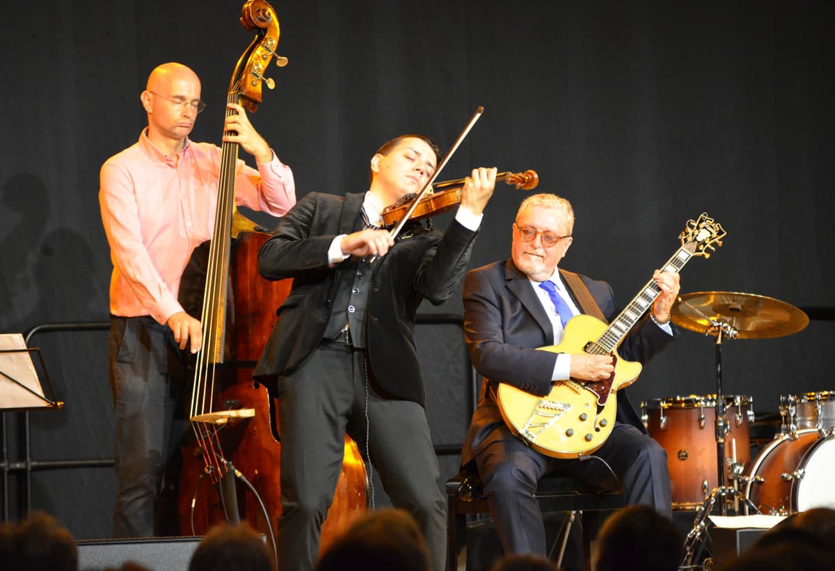 Drei Musikerinnen oder Musiker stehen bzw. sitzen auf einer Bühne und spielen verschiedene Instrumente: ein Kontrabass, eine Violine und eine E-Gitarre. Im Hintergrund ist ein Teil eines Schlagzeugs zu sehen. Die Szene zeigt eine Live-Musikdarbietung mit