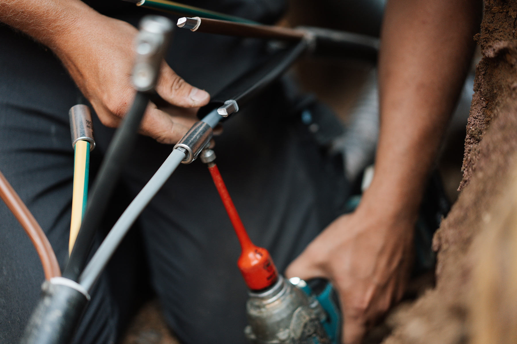 Nahaufnahme der Hände einer Person, die im Außenbereich mit elektrischen Kabeln arbeitet. Eine Hand hält ein Werkzeug mit rotem Griff, die andere Hand justiert oder befestigt mehrere Kabel. Rechts im Bild ist Erde sichtbar, was auf eine Installation oder 