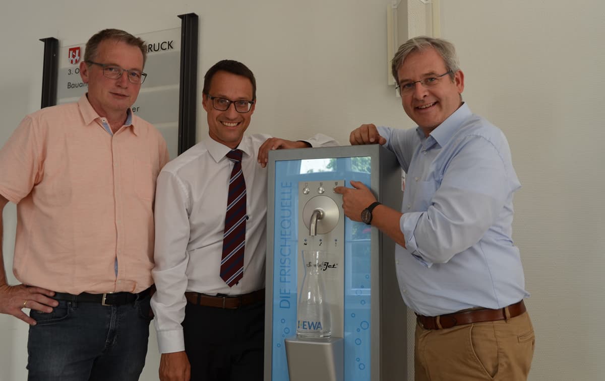 Drei Personen stehen um einen Wasserspender der HEWA GmbH. Die Person links trägt ein rosa Hemd und Jeans, die mittlere Person ein weißes Hemd mit Krawatte, und die rechte Person ein hellblaues Hemd mit brauner Hose. Der Wasserspender steht vor einem blau